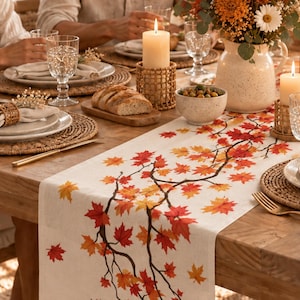 May include: A rectangular off-white table runner featuring a printed pattern of autumn maple leaves in shades of red, orange, and yellow on dark brown branches. The runner is laid across a wooden dining table set with woven placemats and candles.