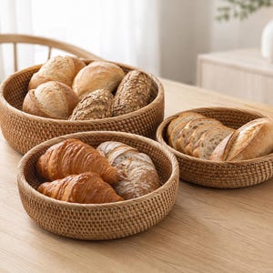May include: Three round, woven rattan baskets in varying sizes, each featuring a natural light brown finish and a tight, uniform weave pattern. The baskets are filled with an assortment of baked goods, including crusty bread rolls and croissants.