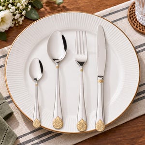 May include: A set of four stainless steel utensils including a small spoon, a large spoon, a fork, and a knife, all featuring a polished silver finish and a decorative gold-toned floral motif at the base of each handle, arranged on a white plate.