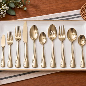 May include: A set of ten gold-toned stainless steel flatware pieces arranged on a rectangular white serving tray with a gold rim. The collection includes various spoons, forks, and a knife, all featuring a smooth, polished finish and a curved handle design.