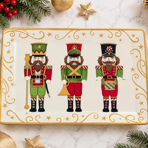 May include: A rectangular white ceramic serving platter featuring a gold-rimmed edge and gold scrollwork with stars. The center displays three illustrated nutcracker figures in red and green uniforms, holding a staff, a horn, and a drum.