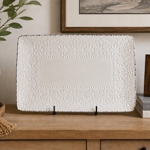 May include: A rectangular white ceramic serving platter featuring an embossed lace-like pattern along the border and a dark, distressed-style rim. The platter is displayed upright on a small black metal stand, showcasing its textured surface.