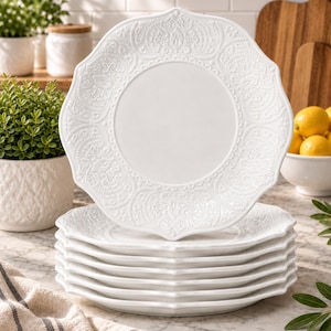 May include: A stack of seven white ceramic plates with a scalloped edge and an intricate embossed floral pattern on the rim. The plates feature a smooth, plain center and a decorative border, presented in a clean, uniform design for dining use.