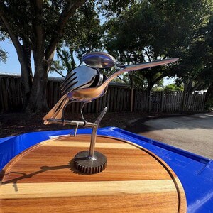 May include: A sculpture of a bird crafted from silver-toned metal cutlery, including spoons and a fork, perched on a metal branch attached to a circular gear base. The bird figure is positioned on a round wooden board with visible grain patterns.