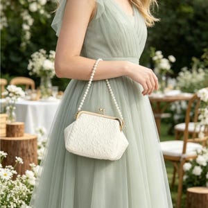 May include: A white clutch bag with a textured surface and a gold-toned metal kiss-lock clasp. The bag features a handle made of a single strand of white pearls. The clutch is held against a sage green dress with a pleated bodice and tulle fabric.