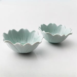 以下が含まれることがあります： 淡いミントグリーンの陶器製ボウル2つ。咲き誇る蓮の花のような形状をしています。各ボウルには波打つような縁取りがあり、中央の底から縁に向かって放射状に伸びるエンボス加工の縦線が施されており、繊細な花びらのような外観を作り出しています。