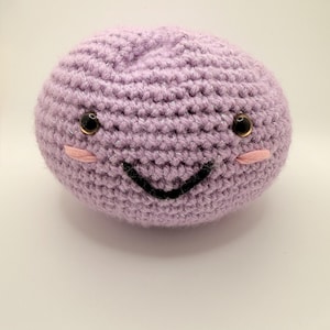 May include: A round, lavender-colored crochet plush toy featuring a smiling face. The toy has two dark brown plastic safety eyes, a stitched black curved mouth, and two small pink embroidered horizontal lines beneath the eyes to represent cheeks.