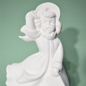 May include: White ceramic figurine of a girl wearing a hat and coat, holding a book. The figurine is in a standing position and has a whimsical expression.