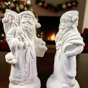 May include: Two unpainted ceramic Santa Claus figurines. One Santa holds a bag and a doll, the other stands with arms crossed. Both figures have long beards, hats, and robes. The figurines are on a dark brown surface, with a Christmas tree and fireplace in the background.