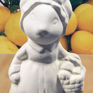 May include: A white ceramic figurine of a character wearing a bonnet and holding a basket. The figurine has a whimsical design and is set against a backdrop of lemons and foliage, suggesting a garden theme.