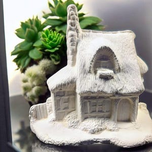 May include: A white ceramic miniature cottage with a thatched roof, chimney, and small windows. The cottage is set in front of a cluster of green succulents, creating a charming, rustic scene. Ideal for home decor.