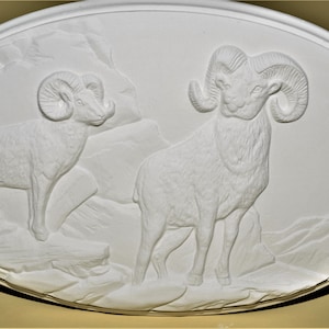 May include: White ceramic wall plaque with a raised relief design of two bighorn sheep standing on a rocky mountain landscape.