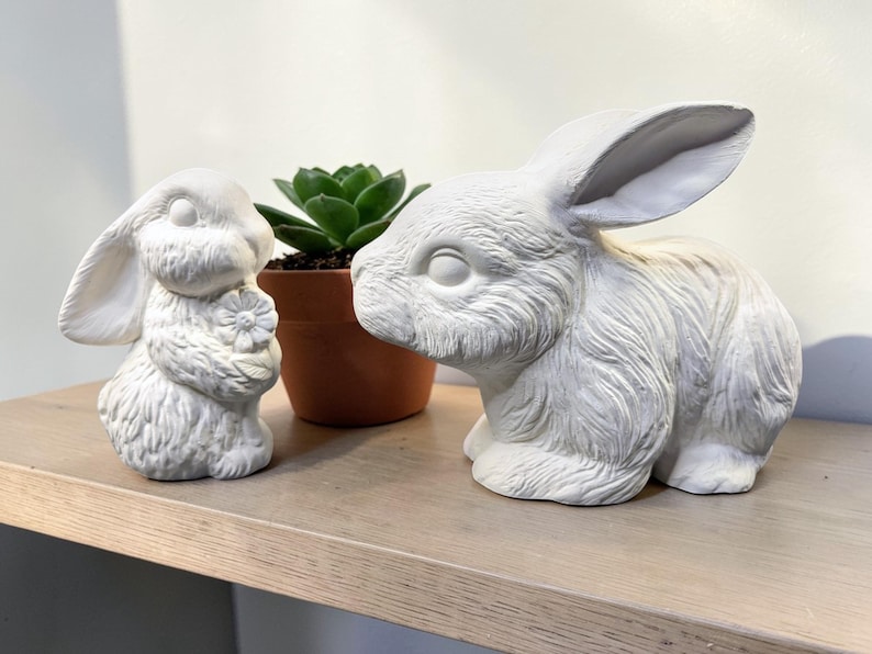 May include: Two white ceramic bunny figurines, one holding a flower, sit on a wooden shelf. A small potted succulent plant is between the bunnies. The bunnies have detailed fur textures and are in a neutral color.