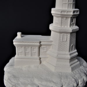 May include: White ceramic lighthouse figurine with a detailed brick exterior and a small chimney on the roof. The lighthouse is perched on a rocky base.
