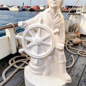 May include: A white statue of a fisherman holding a ship's wheel. The statue is on a wooden deck with ropes and a harbor in the background. The base of the statue reads "Gloucester Fisherman".