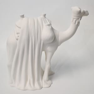 May include: White ceramic camel figurine with a saddle and draped cloth. The camel is standing with its head turned to the right.