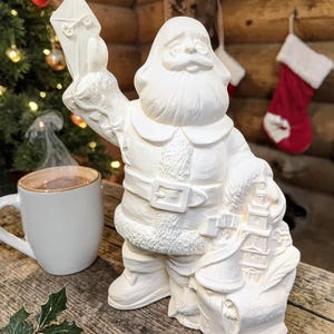 May include: A white ceramic Santa Claus figurine holding a letter and a sack. The figurine has a long white beard, a belt, and a fur-trimmed coat. A mug of coffee sits on a wooden table next to the figurine.