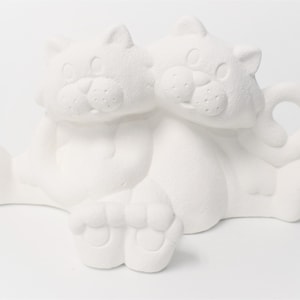 May include: White ceramic figurine of two cats sitting together. The cats are facing forward with their heads touching. The figurine is unpainted and ready to be decorated.