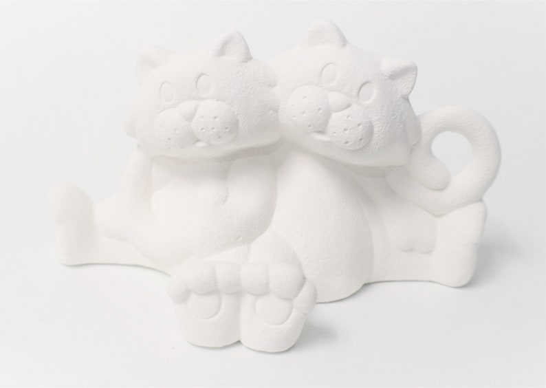 May include: White ceramic figurine of two cats sitting together. The cats are facing forward with their heads touching. The figurine is unpainted and ready to be decorated.