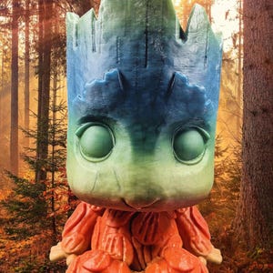 May include: A stylized figure of a tree-like creature with a large head and large eyes. The head is green and blue, and the body is orange. The figure is set against a blurred forest background.