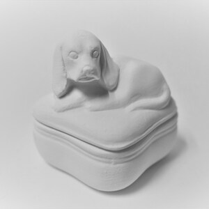 K&ouml;nnte beinhalten: Wei&szlig;e Keramik-Schmuckschatulle mit einem modellierten Hund auf der Oberseite. Die Schachtel hat einen Klappdeckel und eine quadratische Form.