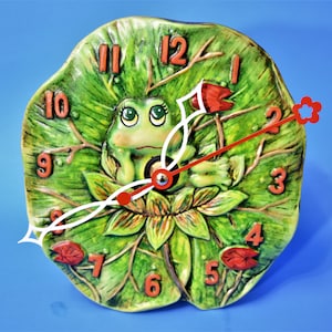May include: A green frog clock with red numbers and hands, sitting on a green lily pad with red flowers. The clock is made of ceramic and has a whimsical design.