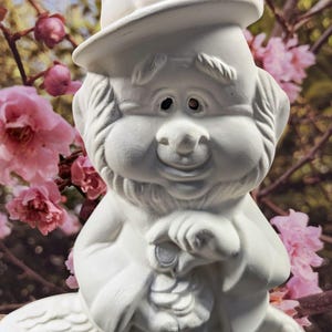 May include: White ceramic figurine of a leprechaun wearing a hat and holding a pot of gold. The figurine is sitting on a rock.