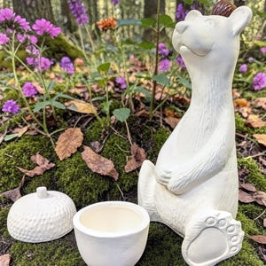May include: A white ceramic bear figurine with a small, round container and a lid. The bear is seated with its paws out, and a small, brown object sits on its head. The objects are on a mossy surface with purple flowers in the background.