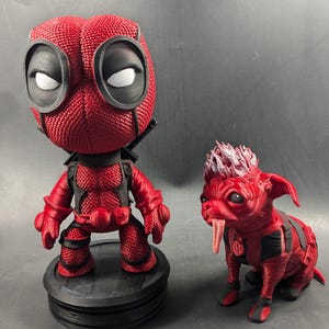 May include: A red and black Deadpool figurine with a red and black dog figurine. Both figurines are standing on a black base.