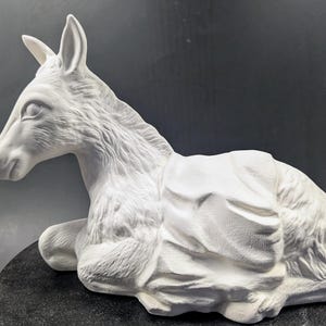 May include: White ceramic figurine of a donkey lying down. The donkey has a saddle and is in a relaxed pose.