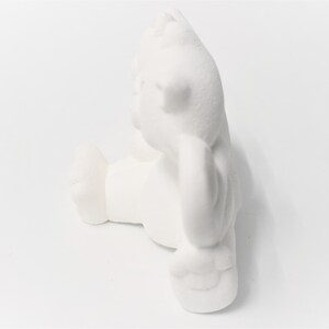 May include: White ceramic figurine of a bear sitting with its paws crossed. The bear is in a relaxed pose with its head tilted slightly to the side.