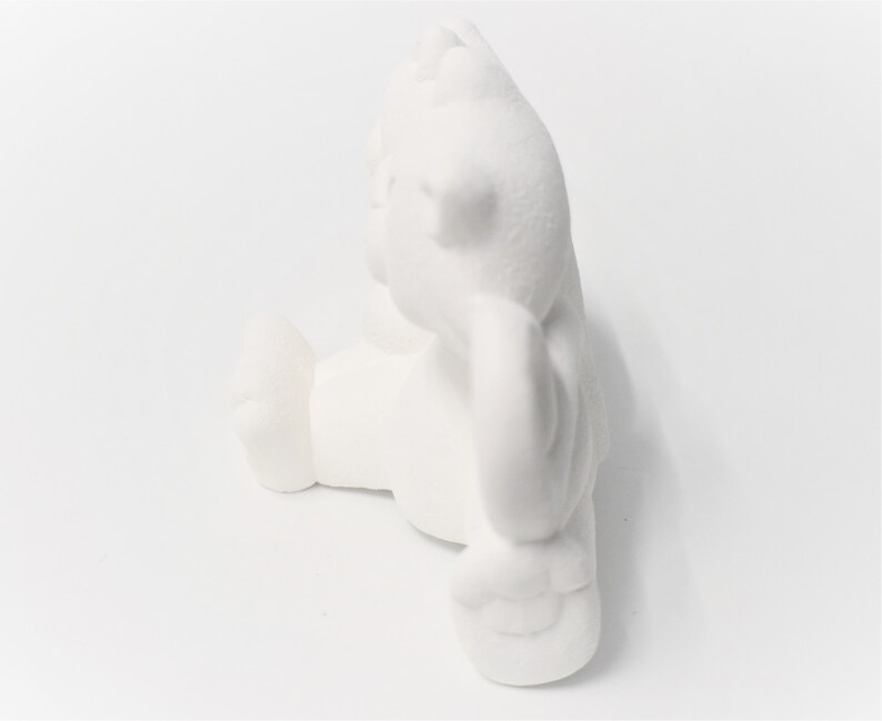 May include: White ceramic figurine of a bear sitting with its paws crossed. The bear is in a relaxed pose with its head tilted slightly to the side.