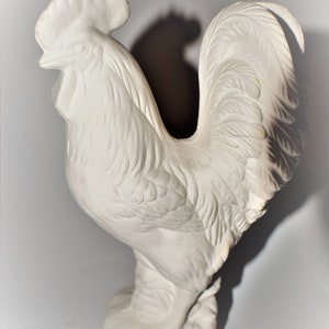 May include: White ceramic rooster figurine with detailed feathers and a raised comb. The rooster is standing on a base with a textured surface.