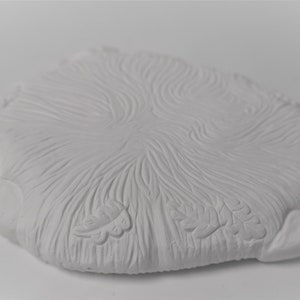 May include: White ceramic coaster with a raised, textured design resembling a tree trunk with leaves. The coaster has a slightly irregular shape.