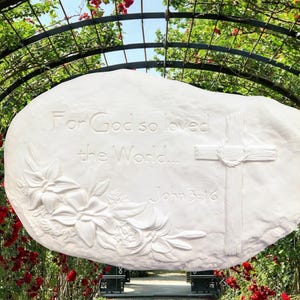 May include: White stone plaque with the inscription "For God so loved the World... John 3:16", a cross, and floral relief. The plaque is set against a backdrop of red roses and greenery.