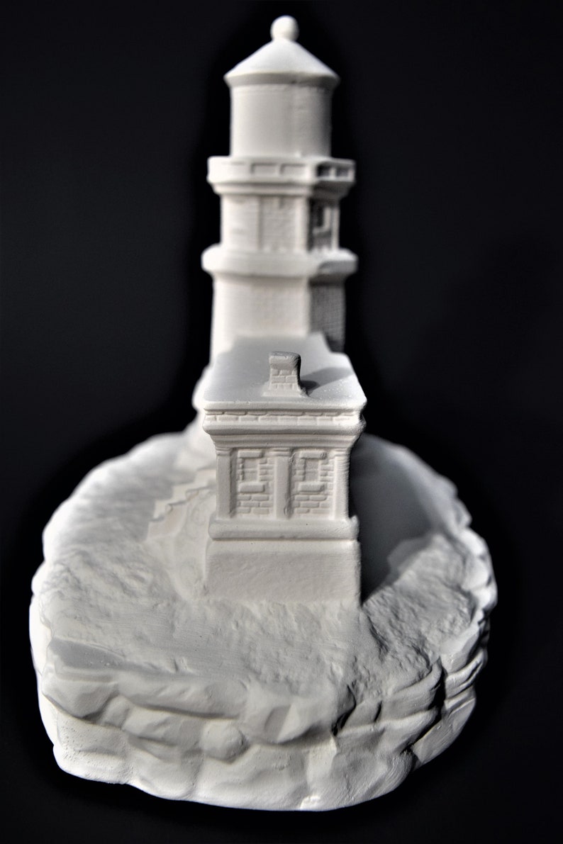 May include: White painted miniature lighthouse figurine with a detailed design, featuring a tower with a small roof and a base that resembles a rocky cliff.