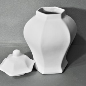 Peut inclure: Un pot en c&eacute;ramique blanc avec un couvercle. Le pot pr&eacute;sente un design g&eacute;om&eacute;trique &agrave; facettes et un corps arrondi. Le couvercle est sur le c&ocirc;t&eacute;, r&eacute;v&eacute;lant l'ouverture du pot. Le fond est gris uni.