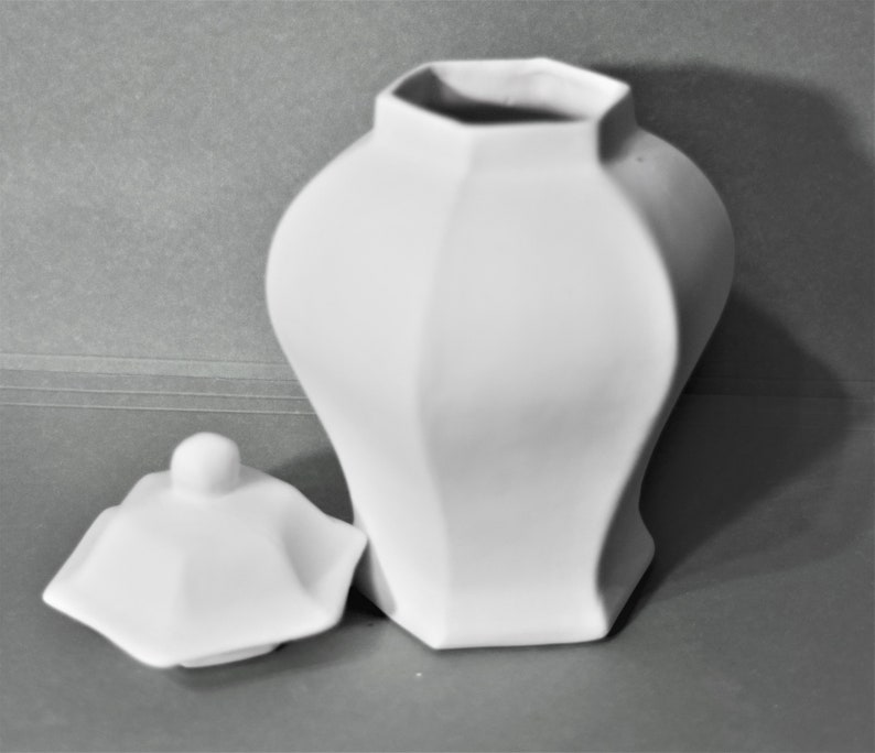 Peut inclure: Un pot en c&eacute;ramique blanc avec un couvercle. Le pot pr&eacute;sente un design g&eacute;om&eacute;trique &agrave; facettes et un corps arrondi. Le couvercle est sur le c&ocirc;t&eacute;, r&eacute;v&eacute;lant l'ouverture du pot. Le fond est gris uni.