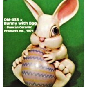 May include: A white ceramic bunny figurine holding a purple and yellow striped egg. The bunny has pink ears and black eyes. The text "DM-435 Bunny with Egg Duncan Ceramic Products Inc. 1971" is printed on the image.