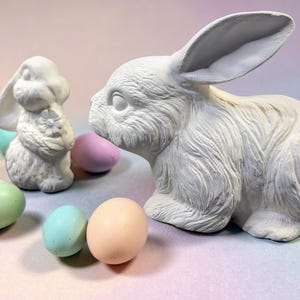May include: Two white ceramic rabbit figurines and several pastel-colored Easter eggs. One rabbit is posed upright, holding a flower, while the other is a larger, seated rabbit. The eggs are in shades of pink, blue, green, and peach.
