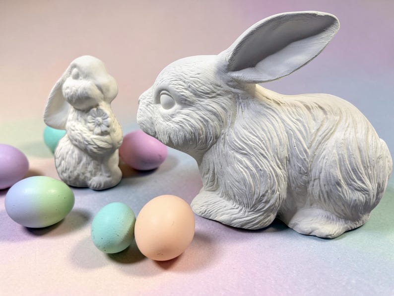 May include: Two white ceramic rabbit figurines and several pastel-colored Easter eggs. One rabbit is posed upright, holding a flower, while the other is a larger, seated rabbit. The eggs are in shades of pink, blue, green, and peach.