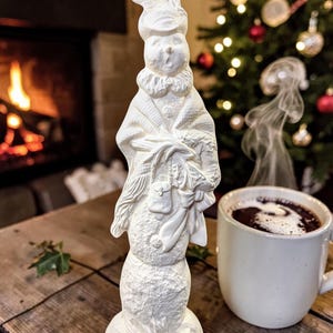 May include: A white ceramic snowman figurine wearing a hat, a ruffled collar, and a shawl, holding a small bag and a bundle of items. The snowman has a textured, snow-like surface and stands upright on a circular base with a detailed finish.