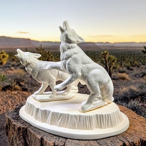 May include: A white ceramic sculpture of two wolves howling. One wolf stands on its hind legs, while the other is on all fours. The sculpture is set on a white base, and the background features a desert landscape.