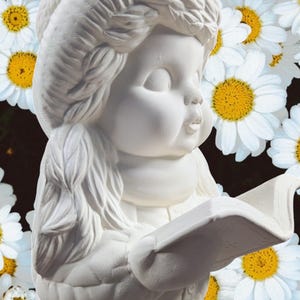 May include: White ceramic figurine of a person holding a book. The person has a braided hairstyle and is wearing a long-sleeved shirt and pants. The figurine is surrounded by white daisies.