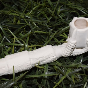 May include: White ceramic pipe shaped like a dinosaur claw. The pipe is unpainted and ready to be decorated.