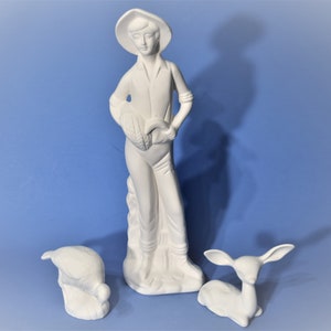 May include: White ceramic figurine set of a person holding a basket, a bird, and a deer. The person is wearing a hat and has a long dress. The bird is sitting on the ground and the deer is standing on its hind legs.