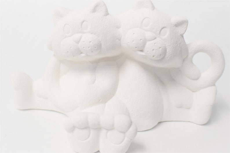 May include: White ceramic figurine of two cats sitting together. The cats are facing forward with their heads touching. The figurine is unpainted and ready to be decorated.
