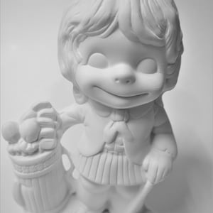 May include: White ceramic figurine of a girl wearing a dress and a bow in her hair. She is holding a golf bag with golf balls.