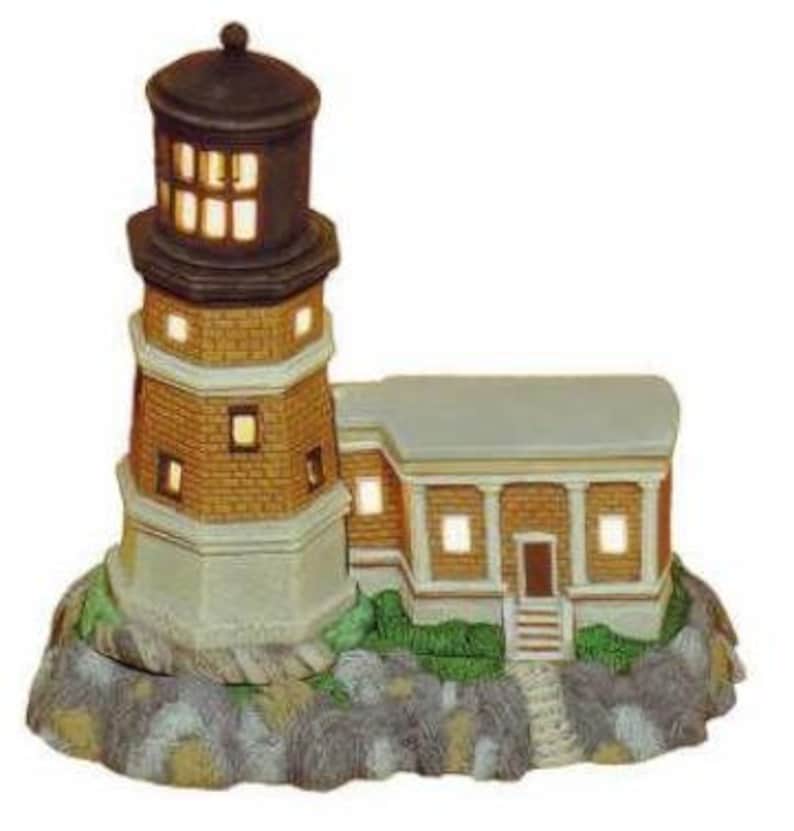 May include: A miniature ceramic lighthouse with a brown roof and brick exterior, illuminated with warm white lights. The lighthouse is set on a base with green grass and a stone path.