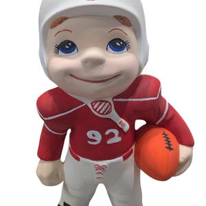 May include: A ceramic figurine of a football player wearing a red and white uniform with the number 92 on the front. The player is holding a brown football and has a white helmet with a red stripe.